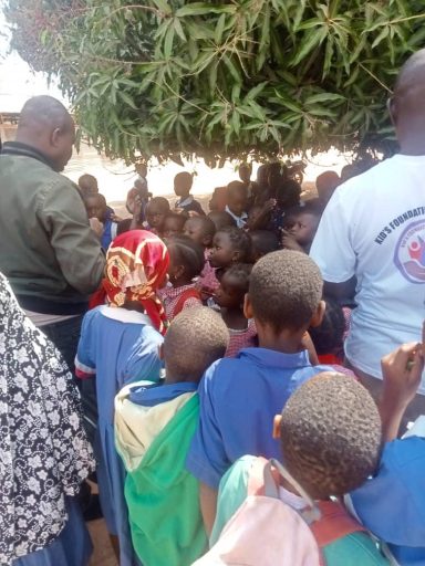 Group of children gathered under a tree, listening attentively to an adult speaker.